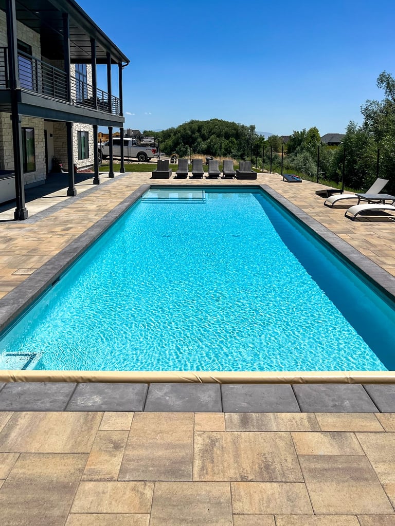 A bright blue rectangular pool with surrounding stone patio, modern two-story home, lounge chairs, and landscaped grounds under clear sky