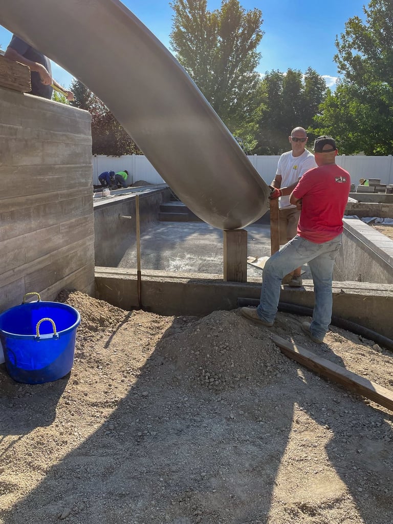 Workers pouring concrete from a large metal chute into a foundation form during construction, with a blue bucket nearby