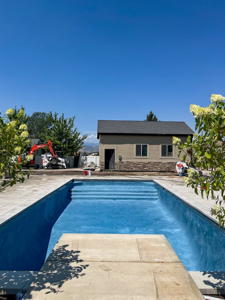 Empty residential swimming pool with concrete deck, house and flowering plants under clear blue sky