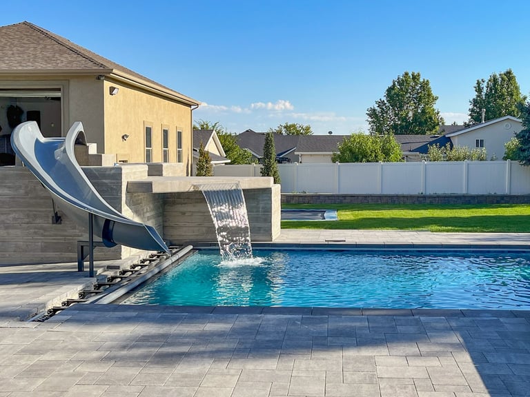 Residential backyard with a swimming pool and metal slide attached to a poolhouse, surrounded by a fence and landscaped lawn