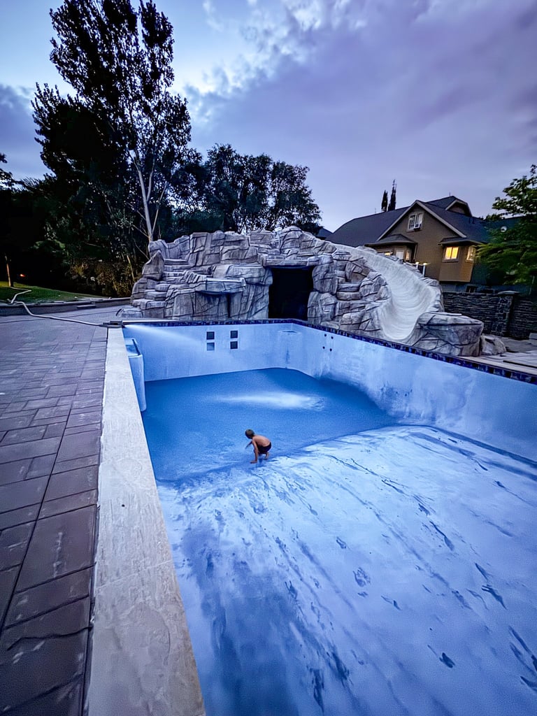 Residential backyard with an empty swimming pool under construction and a stone waterslide structure at dusk