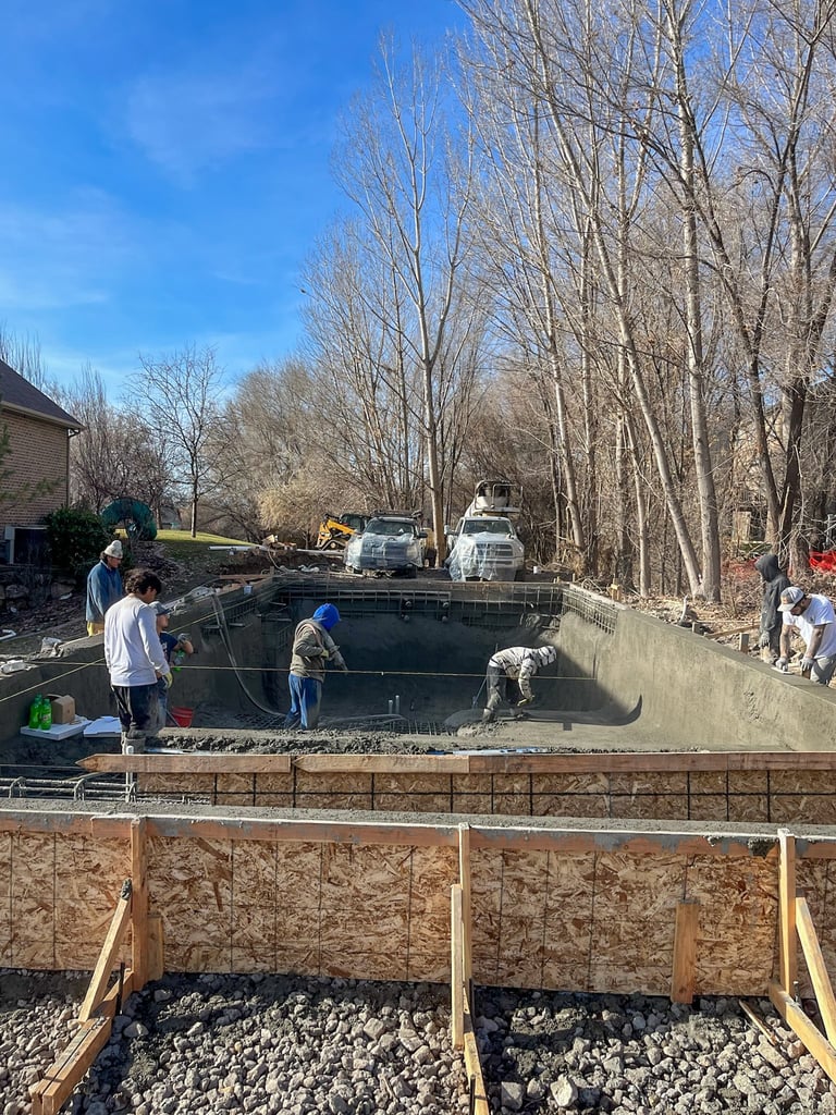 Construction workers pouring concrete into a large rectangular foundation pit with bare trees and residential buildings in the background on a clear day