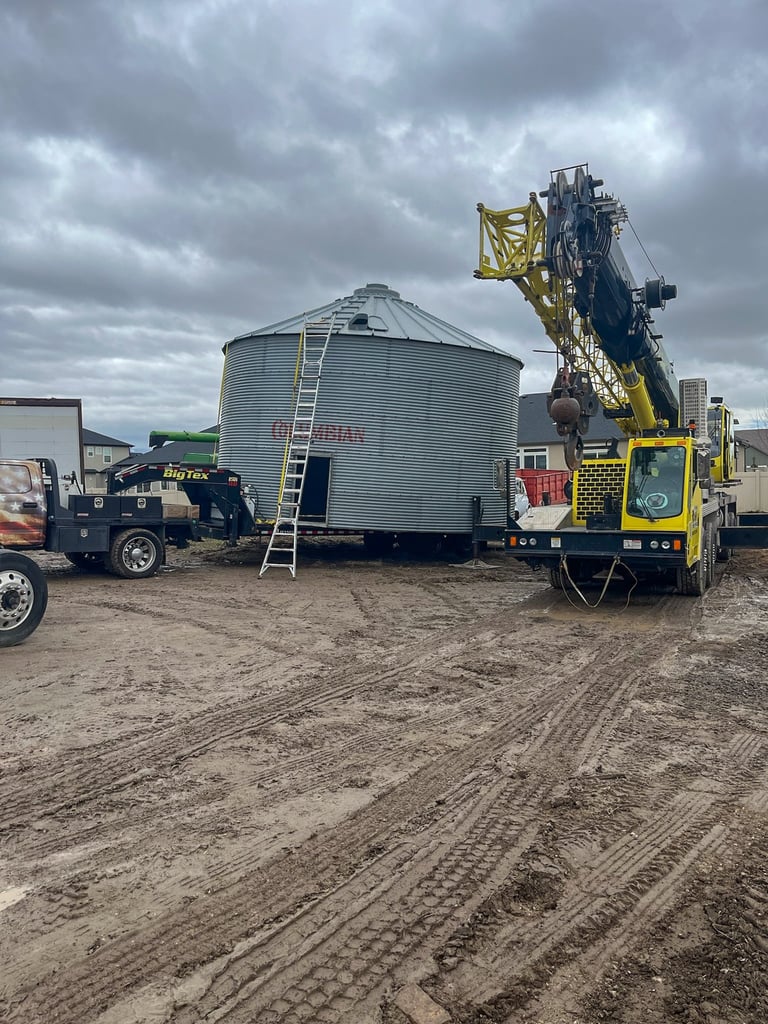 Yellow grain auger truck unloading into a large metal grain bin on a construction lot under cloudy skies