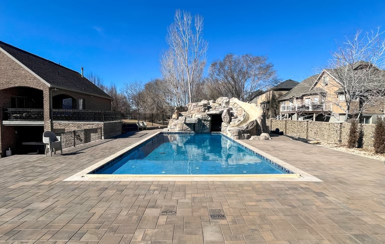 Luxurious backyard with rectangular swimming pool, stone patio, and large brick house under clear blue sky