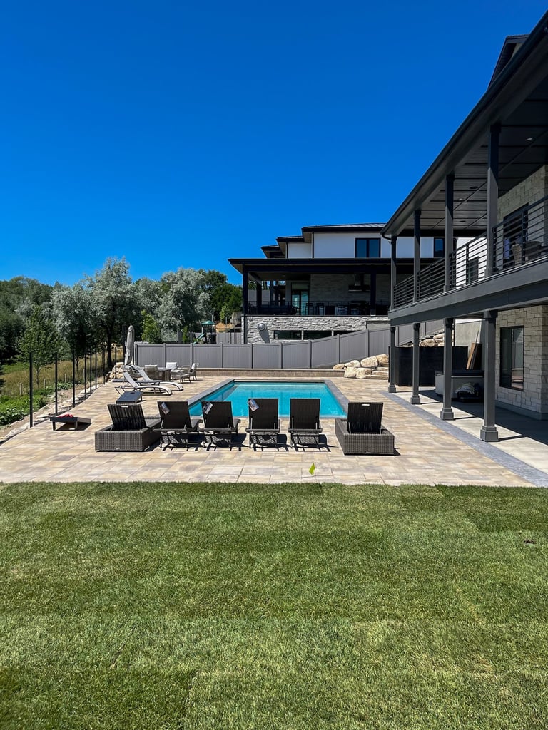 Modern home with rectangular swimming pool, lounge chairs on paved patio, manicured lawn, and trees under clear blue sky