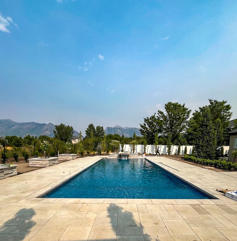 Large rectangular swimming pool with clear blue water surrounded by stone patio, green trees, and mountains in the background under clear blue sky