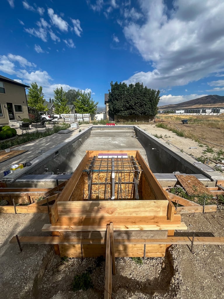 Construction site showing reinforced concrete foundation with wooden forms and rebar framework under clear blue sky with scattered clouds