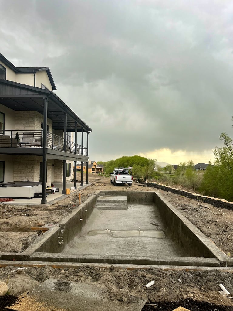 Modern two-story house under construction with pool foundation, work truck, and green landscape under cloudy sky
