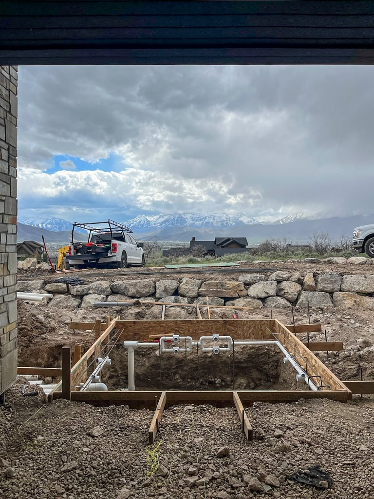 Construction foundation pit with wooden framing and white PVC pipes, vehicles and snow-capped mountains visible in background