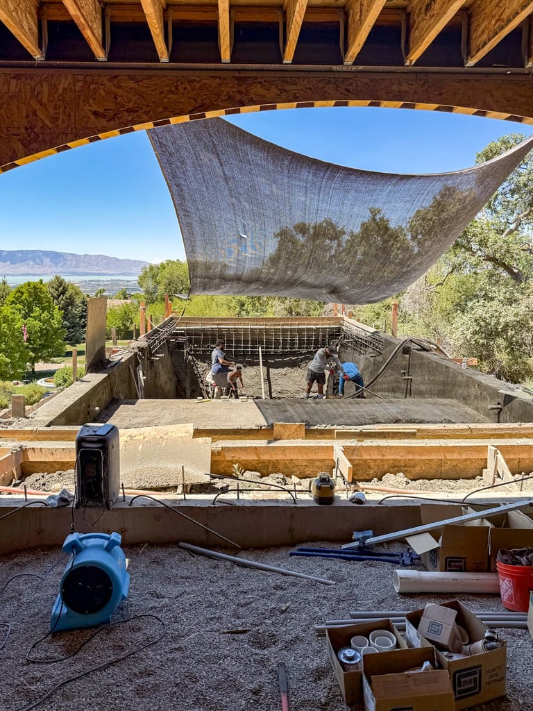 Construction site with wooden frame structure and gray shade cloth overhead, overlooking valley landscape with workers in sunken pit area