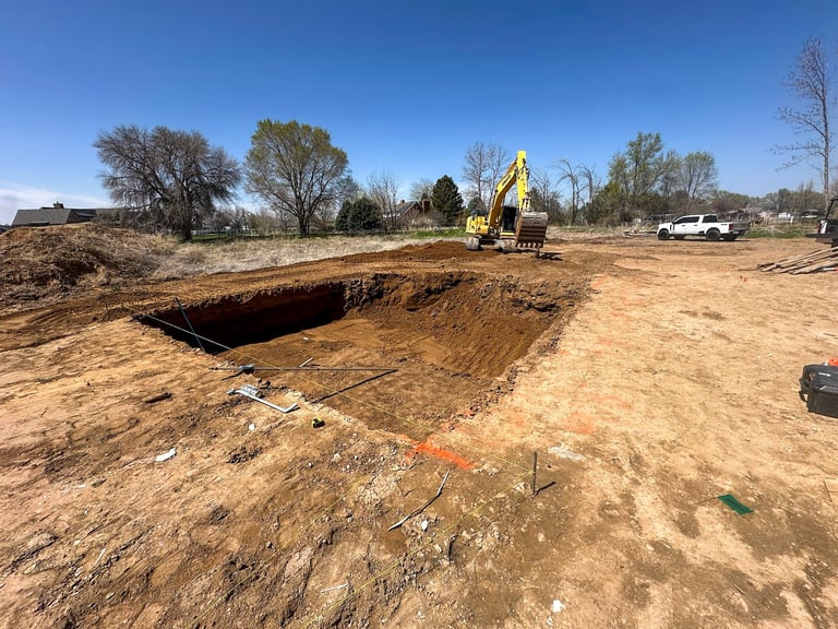 Yellow excavator digging a large hole in a dirt lot with bare trees and white truck visible in the background