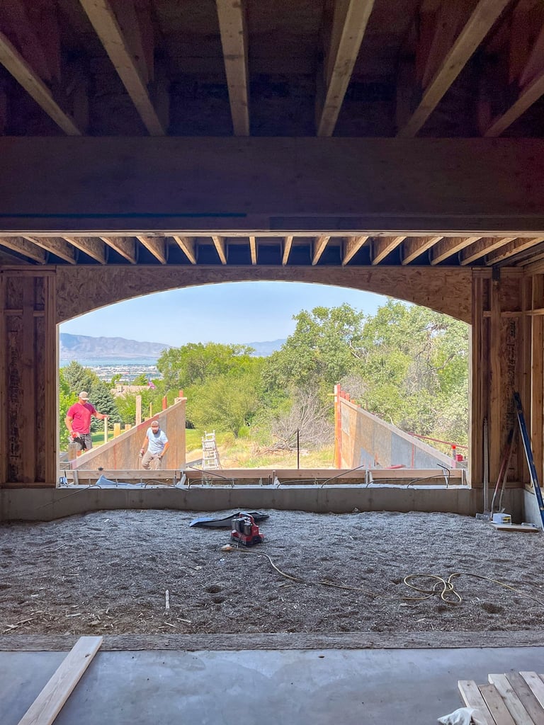 Interior view of a house under construction with exposed framing, showing a scenic mountain and valley landscape through the open front window