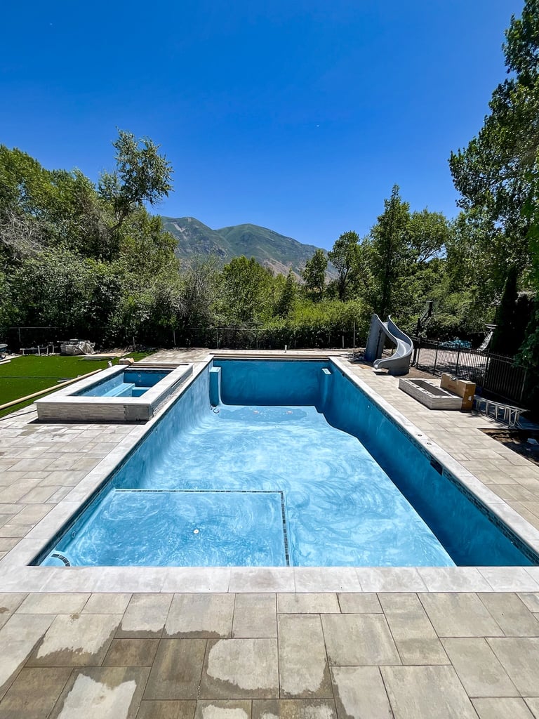 Modern swimming pool with blue water surrounded by stone patio, spa tub, green lawn, and mountains in background under clear sky