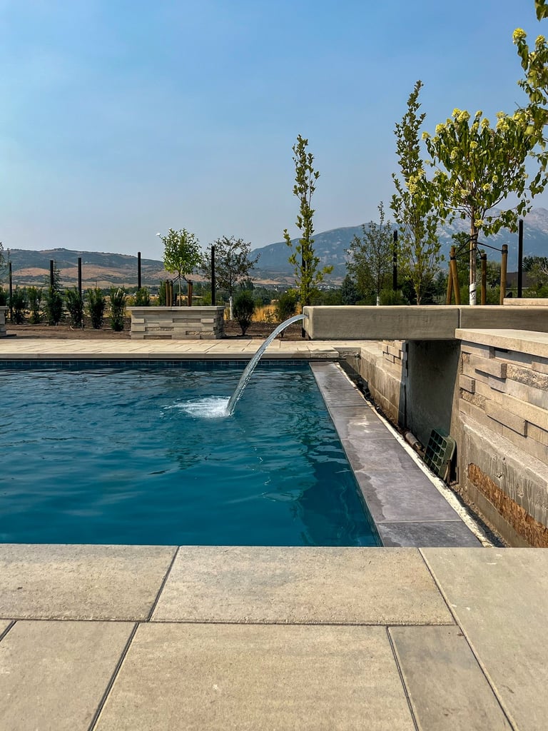 Modern swimming pool with waterfall feature, concrete deck, and mountain landscape with trees in background under clear blue sky