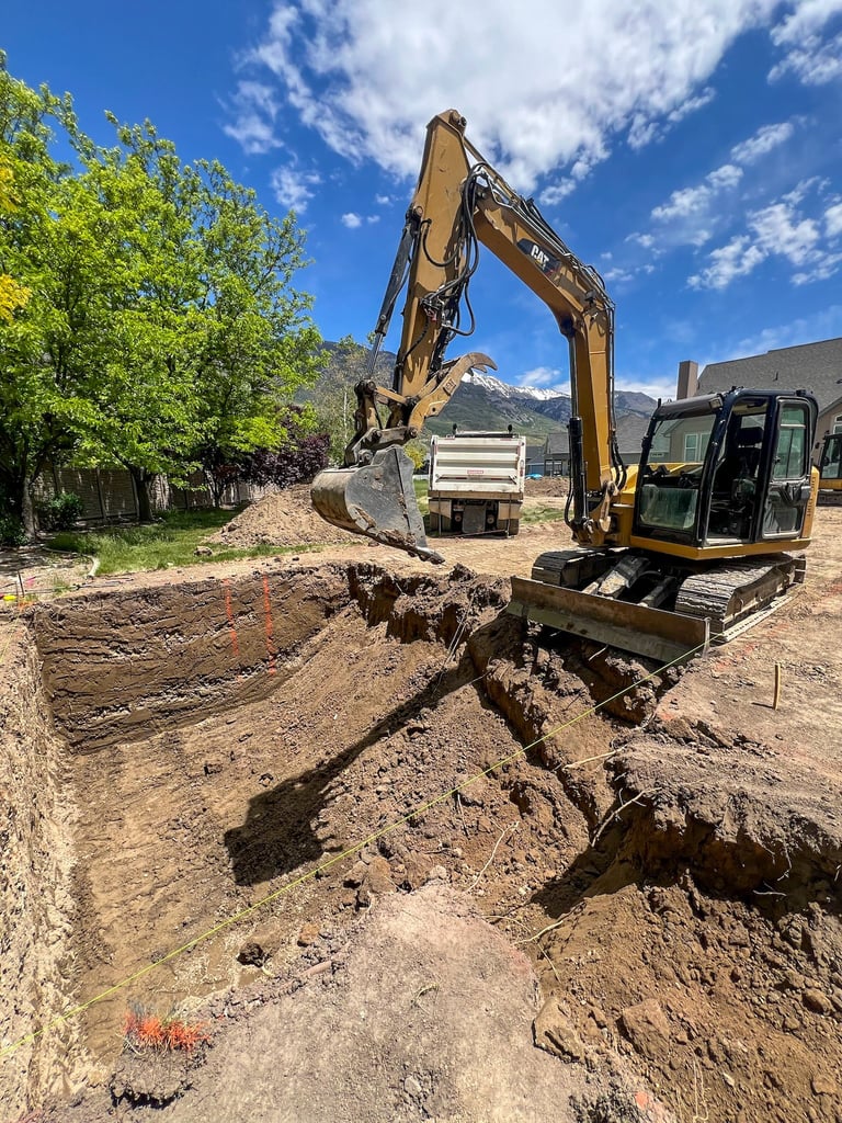 Excavator operating on construction site, digging into soil with bucket raised, mountains visible in background
