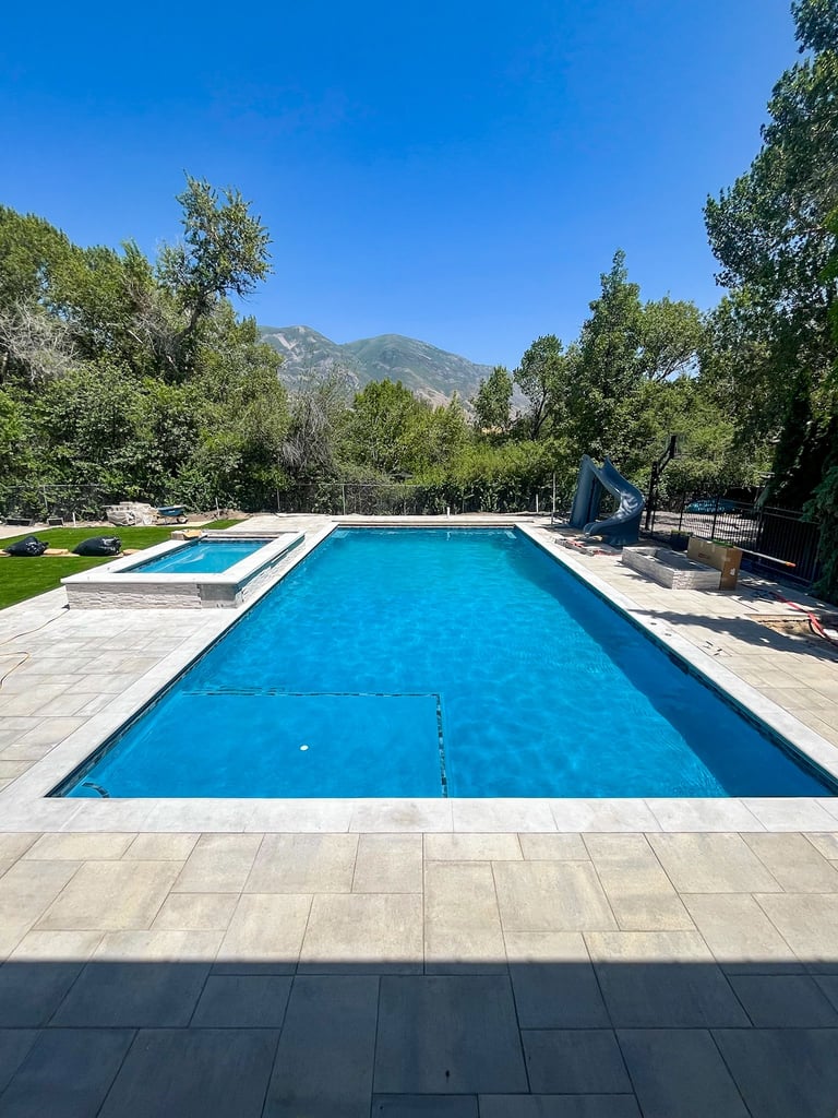 A rectangular swimming pool with bright blue water surrounded by stone patio, manicured lawn, trees, and mountains in the background under clear blue sky