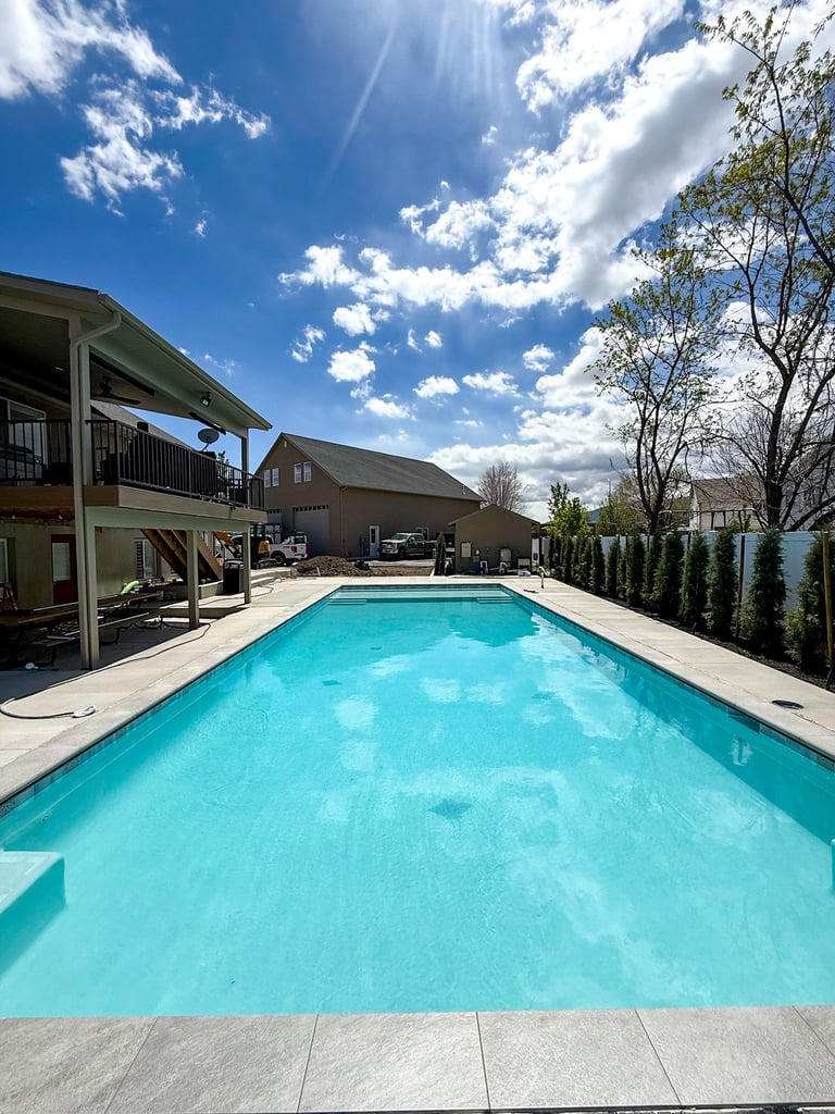 Rectangular swimming pool with turquoise water in a backyard, flanked by modern houses and evergreen trees under blue sky